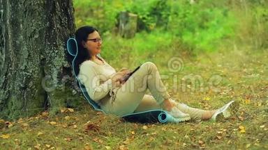 一个戴眼镜的女人<strong>坐在公园里</strong>的树后面，用平板电脑工作